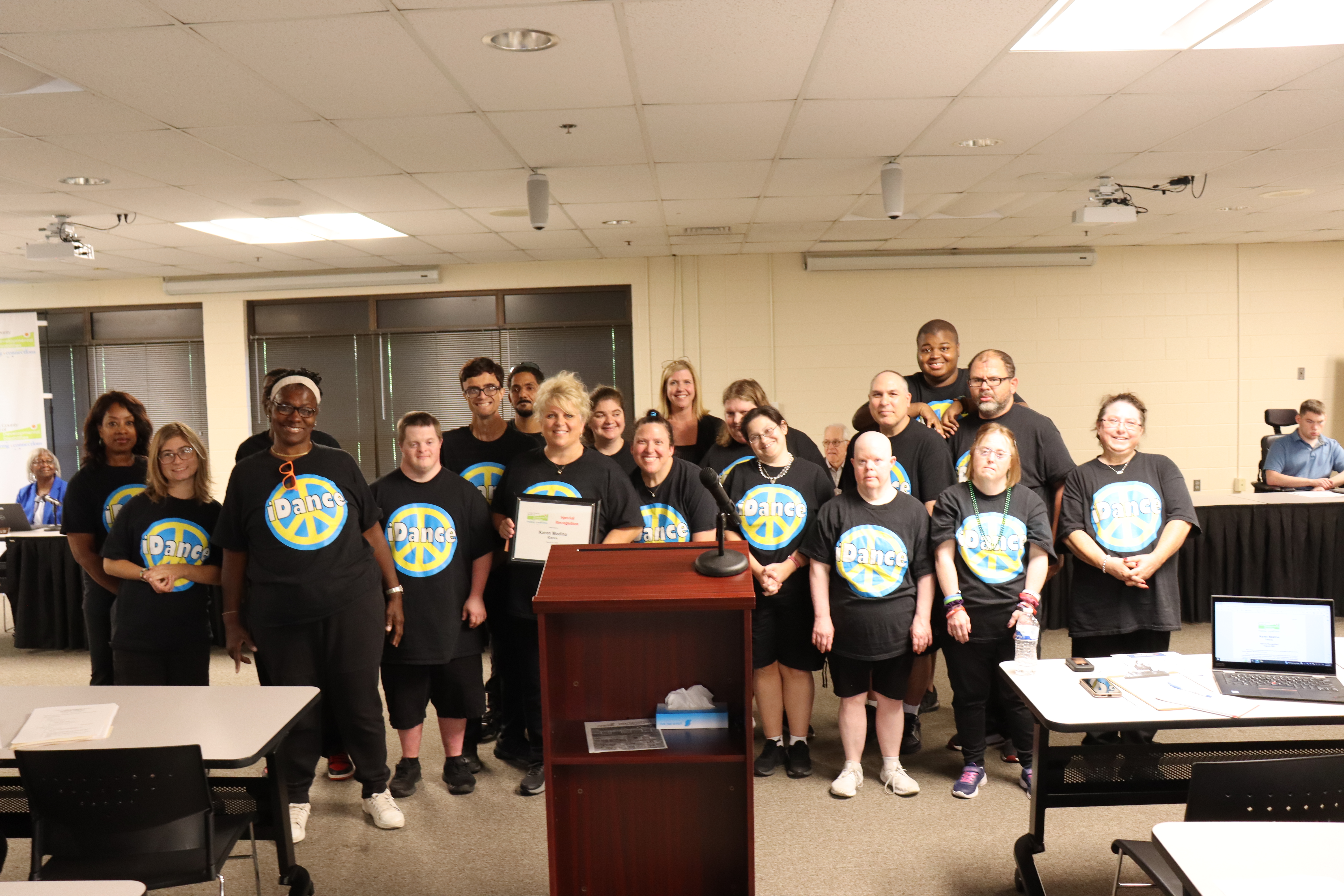 Podium in the foreground with iDance performers surrounding Karen Medina, all wearing black tshirts with a peace symbol and the word iDance on the chest.