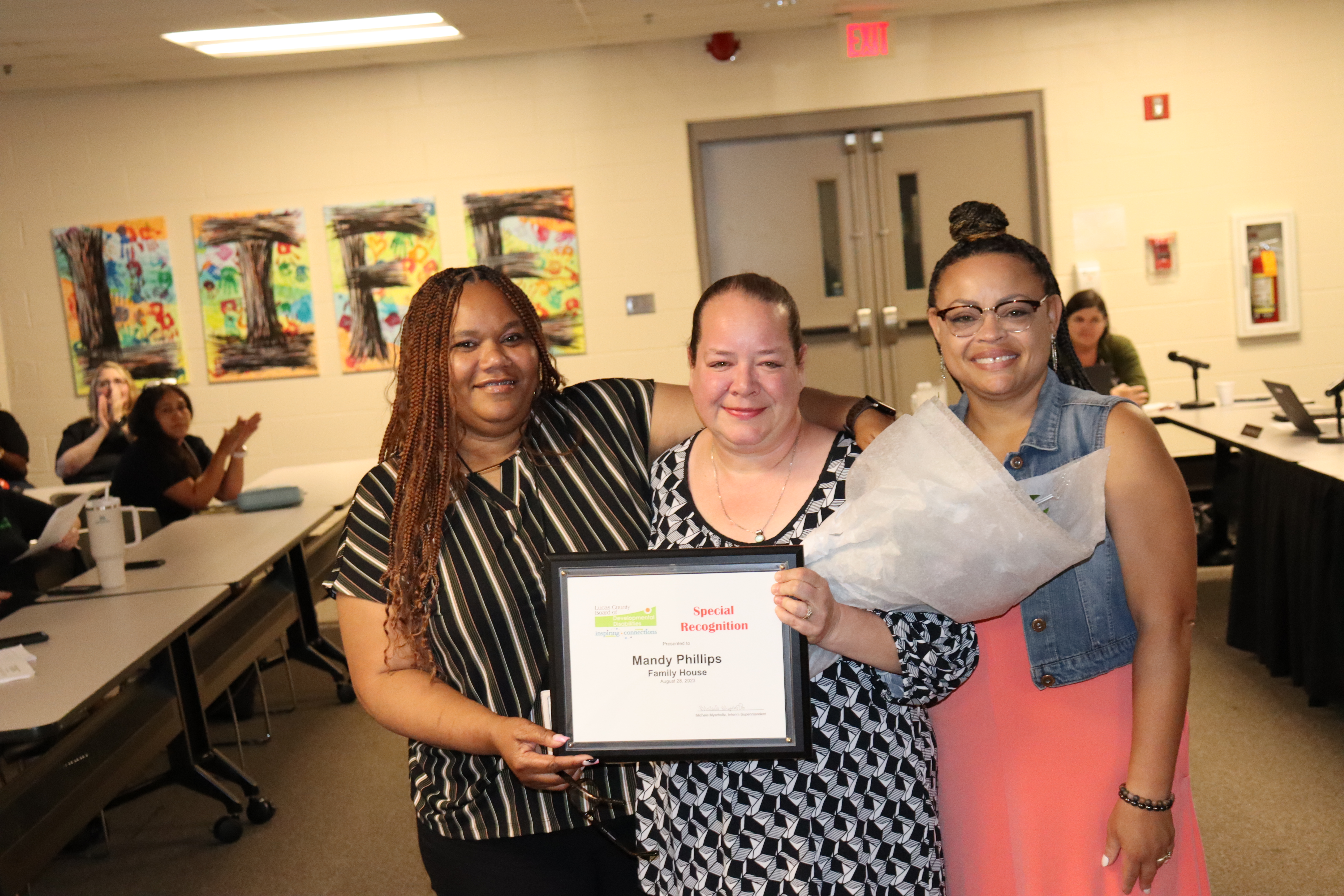 Photo of Mandy Phillips, center, flanked by Lucas DD Self Advocacy & Community Inclusion Specialist Deetra Mitchell on the left, and Community Connections Trainer Sharese Wilburn on the right. Crowd applauding in the background.