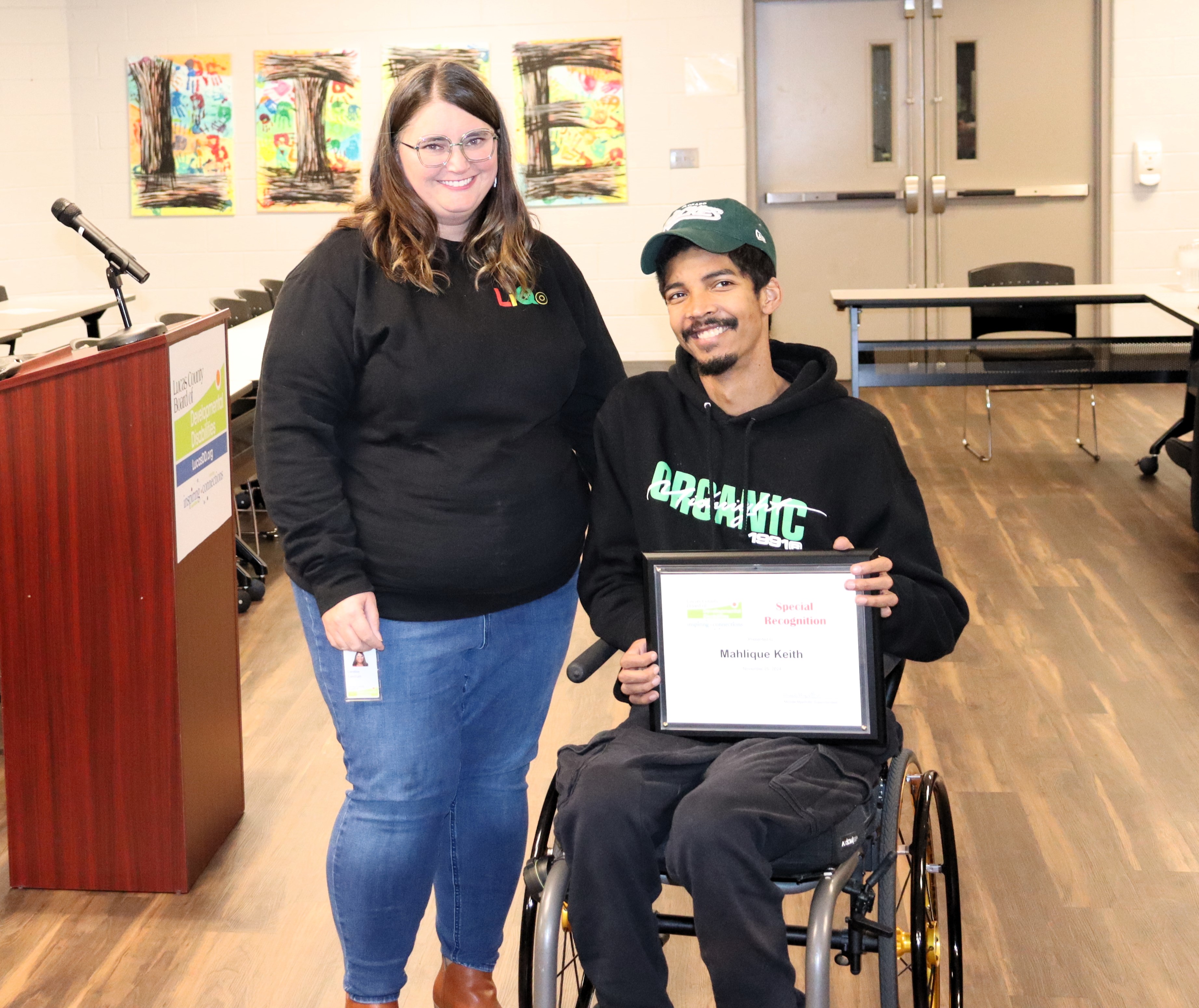 Mahlique Keith on the right in a wheelchair holding his award plaque. He is in a black hoodie and green baseball cap.