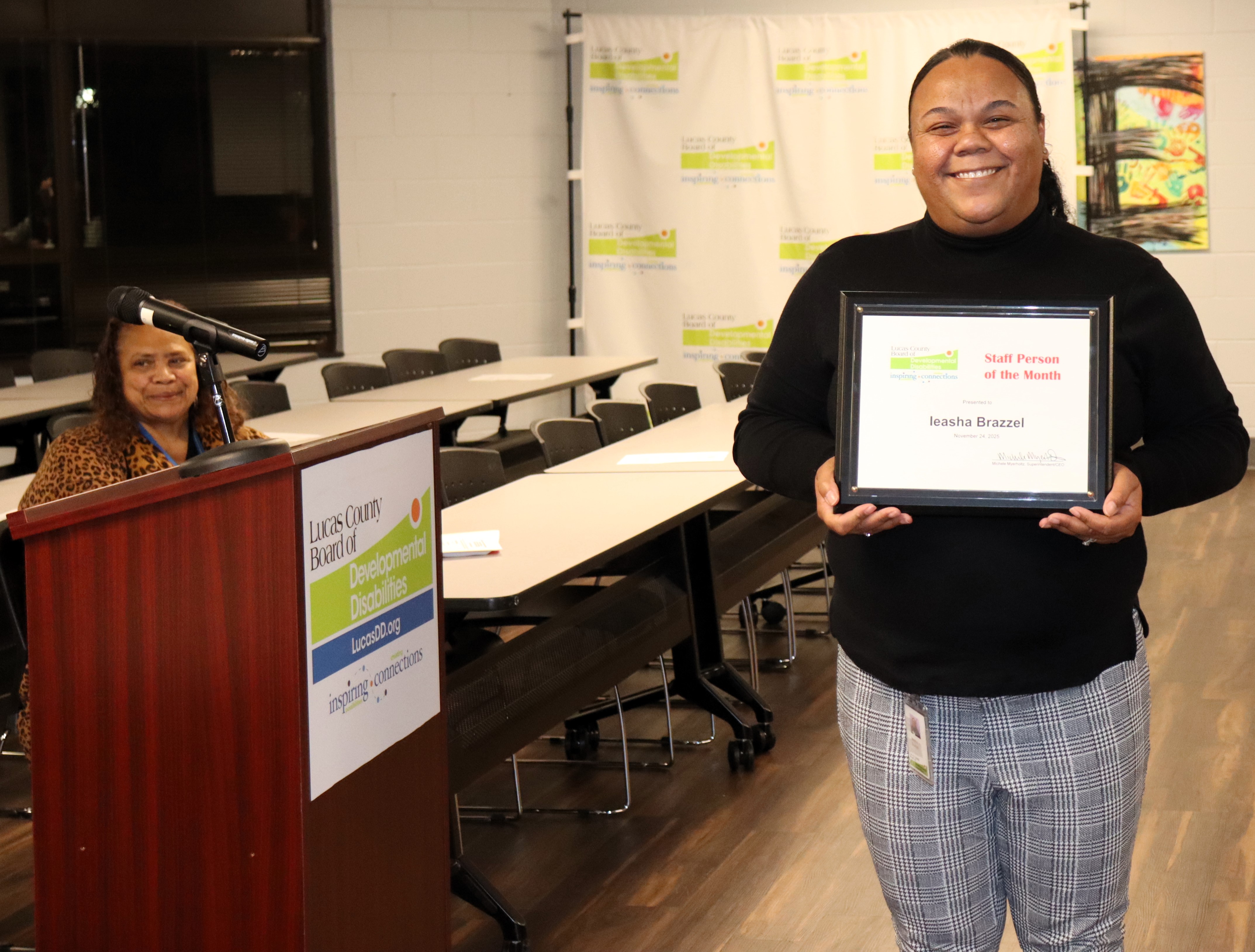 Ieasha holds her plaque as she poses for a photo with the podium to her right and the Lucas DD backdrop behind her.