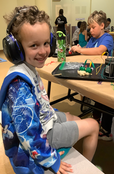 Boy wearing blue noise-cancelling headphones sits at a table during summer camp